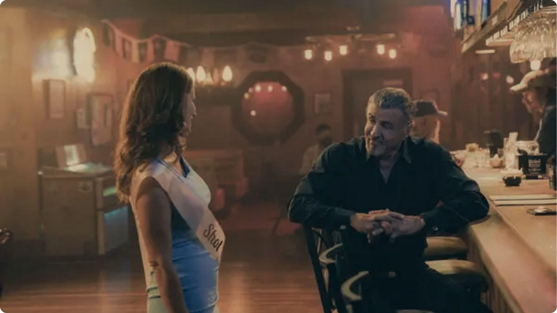 Sylvester Stallone sits at a dim bar talking to a woman with a Shot sash inside the BRED2BUCK Saloon set for Tulsa King.
