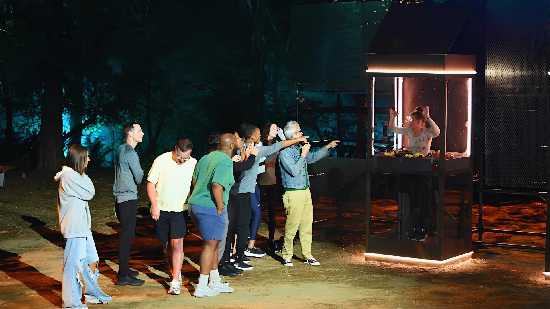 Night scene of group watching woman in lit glass challenge booth filled with bugs host in denim jacket points as contestants react.