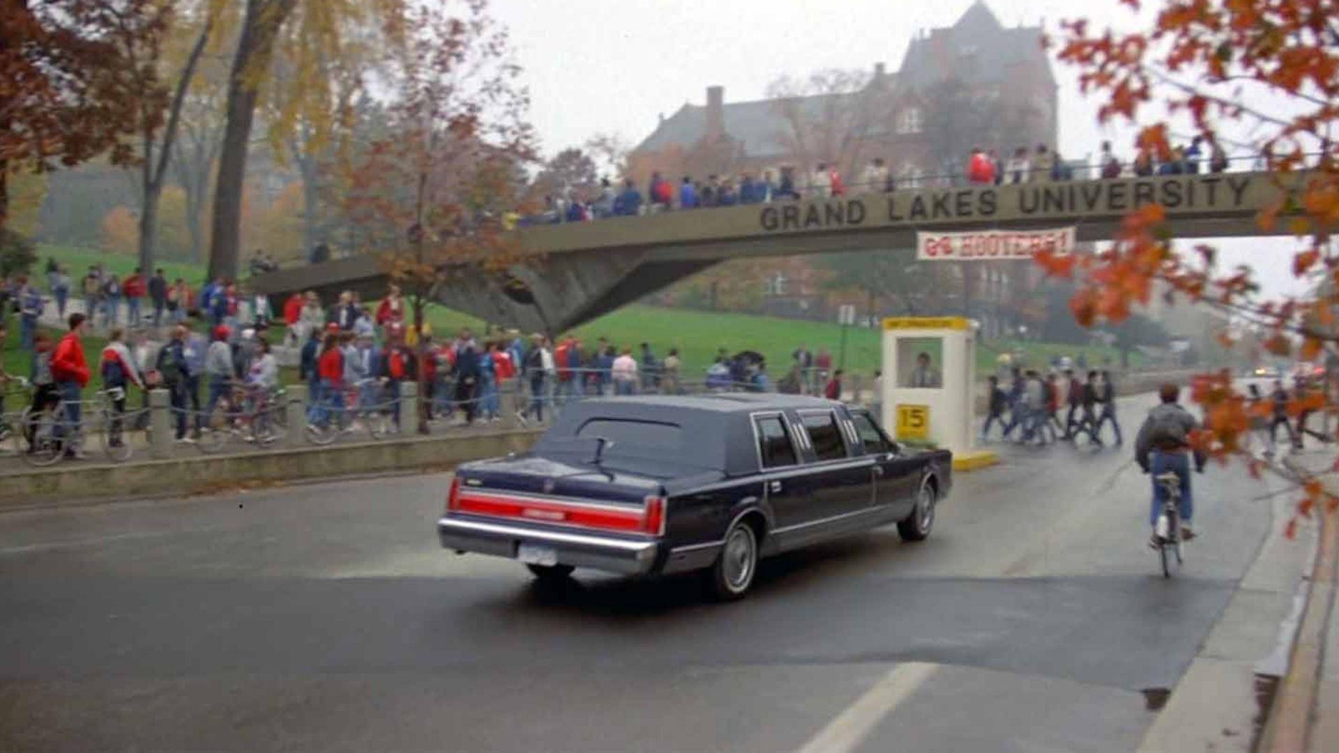 A stretch limousine passes under the Grand Lakes University bridge amid crowds of students in autumn.