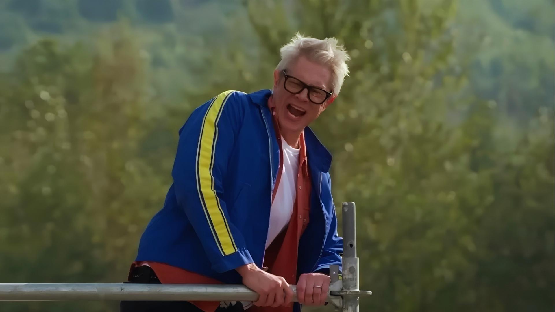 Man with spiky gray hair and black glasses wearing blue track jacket with yellow stripe shouts while gripping metal railing, trees blurred in background.