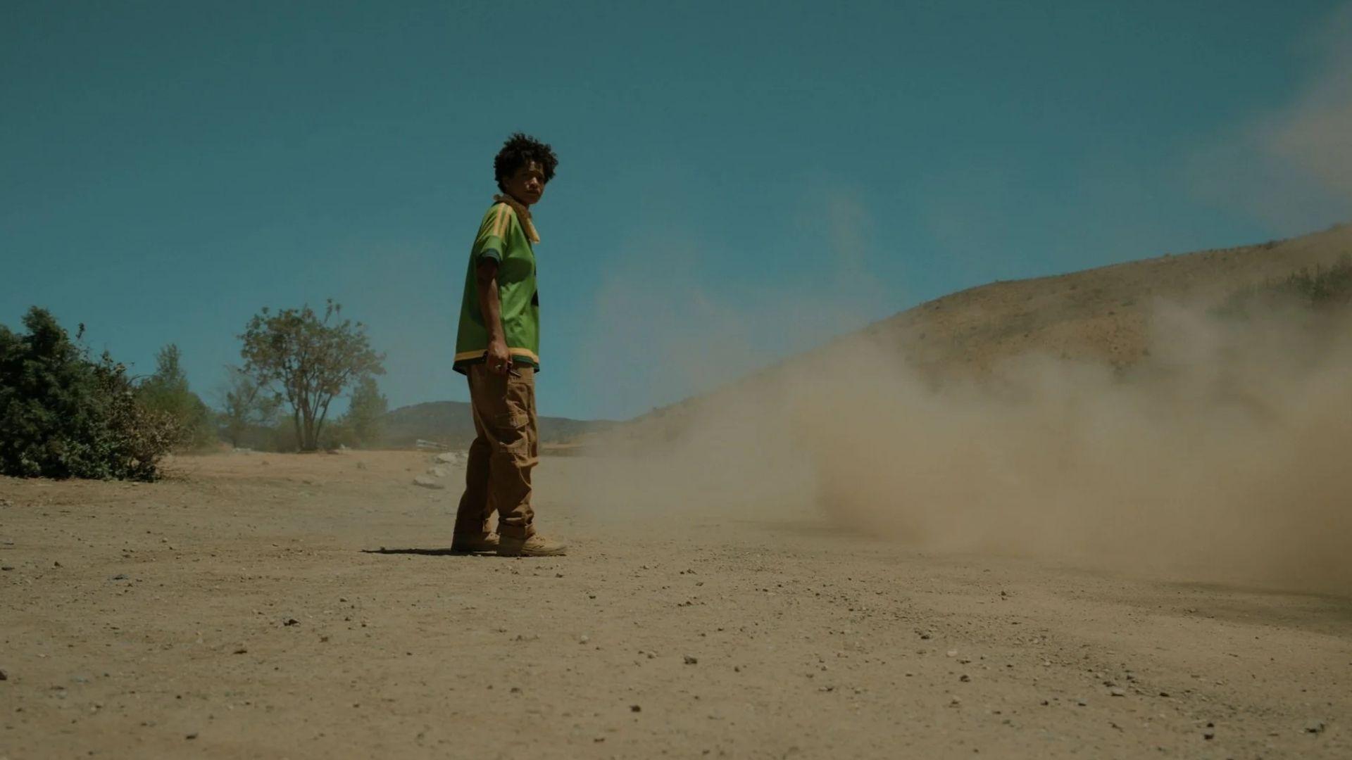 Still from NOPE. Teen Jupe in green polo looks back as a massive dust cloud rises on a barren dirt road.