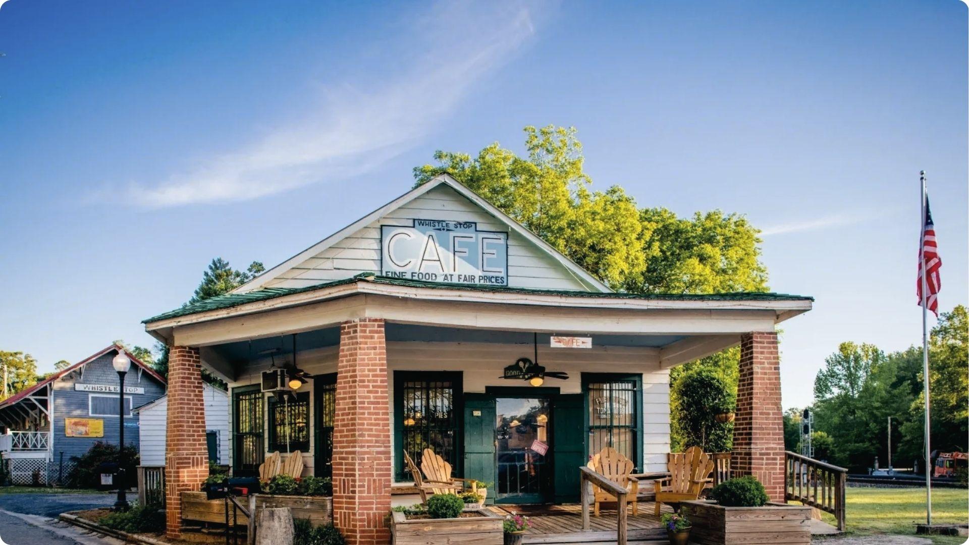 Whistle Stop Cafe exterior with American flag, porch, railroad crossing, fall trees behind.
