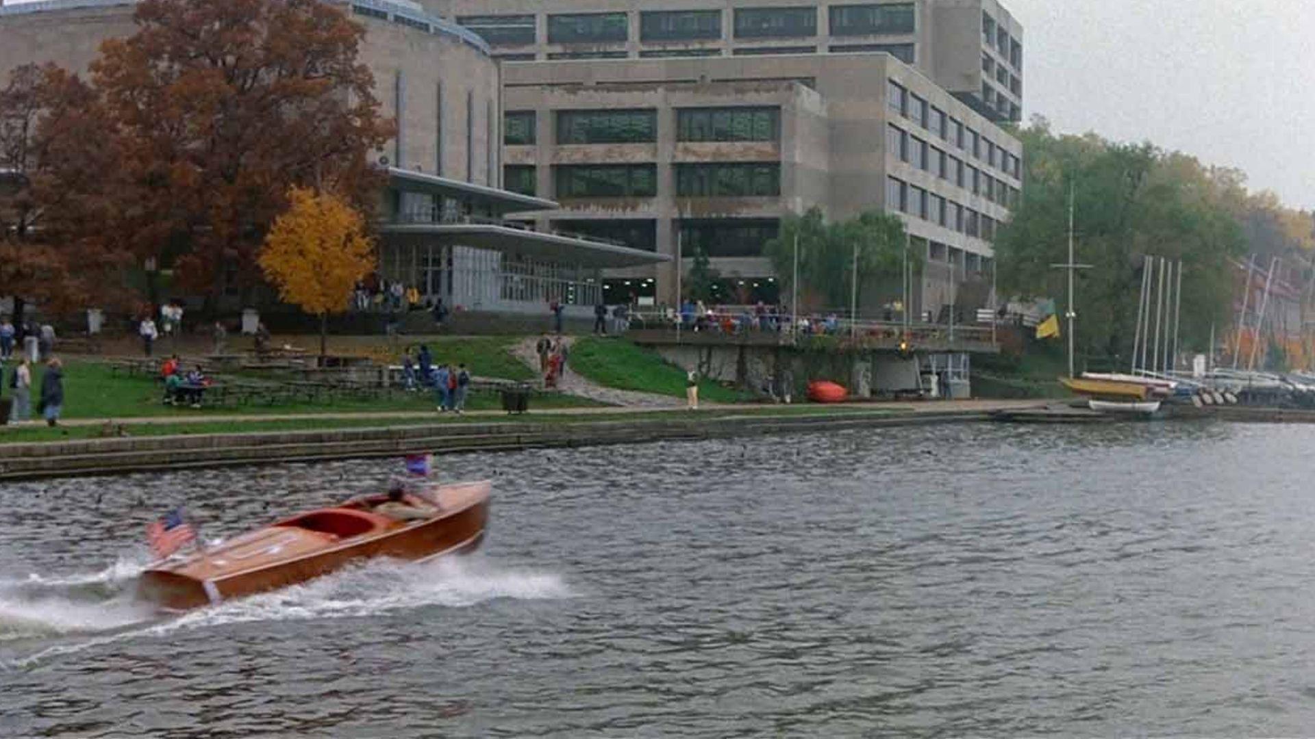 A vintage wooden speedboat races past the university campus building on a grey autumn day.