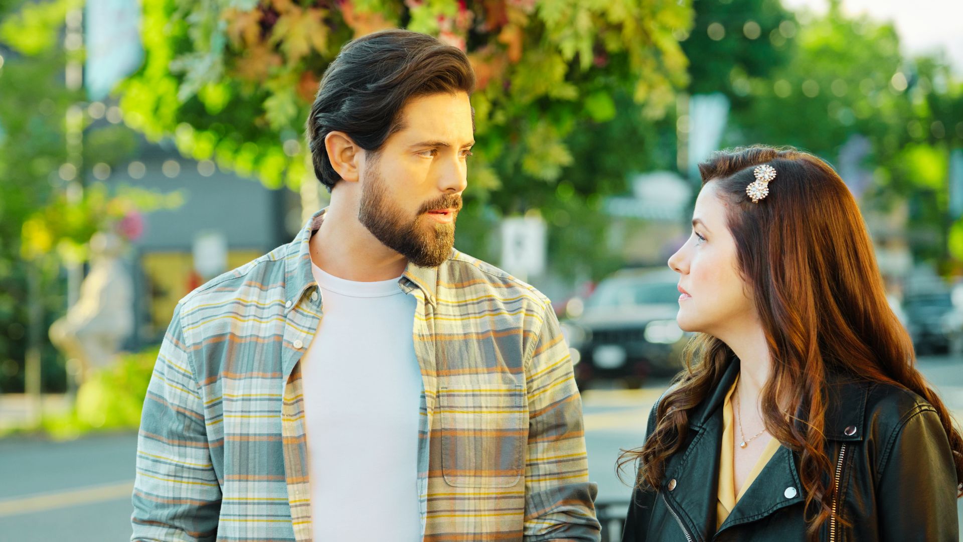 A man in a plaid shirt and woman in a leather jacket share a tense look on a sunny street.