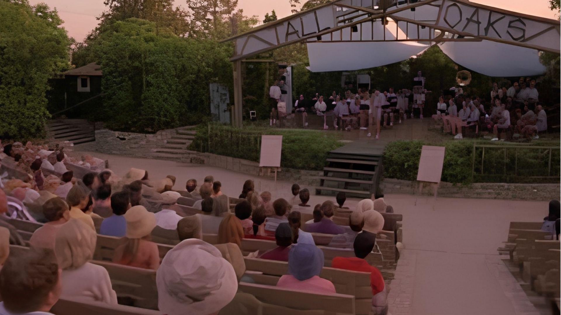 An audience watches a summer evening concert at an outdoor bandshell labelled "All Oaks."