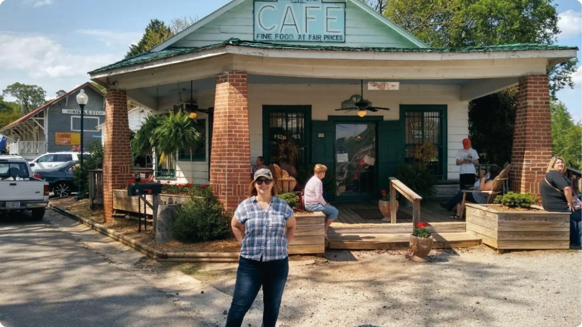 A woman posing in front of Whistle Stop Cafe with a green roof, brick pillars, porch chairs.
