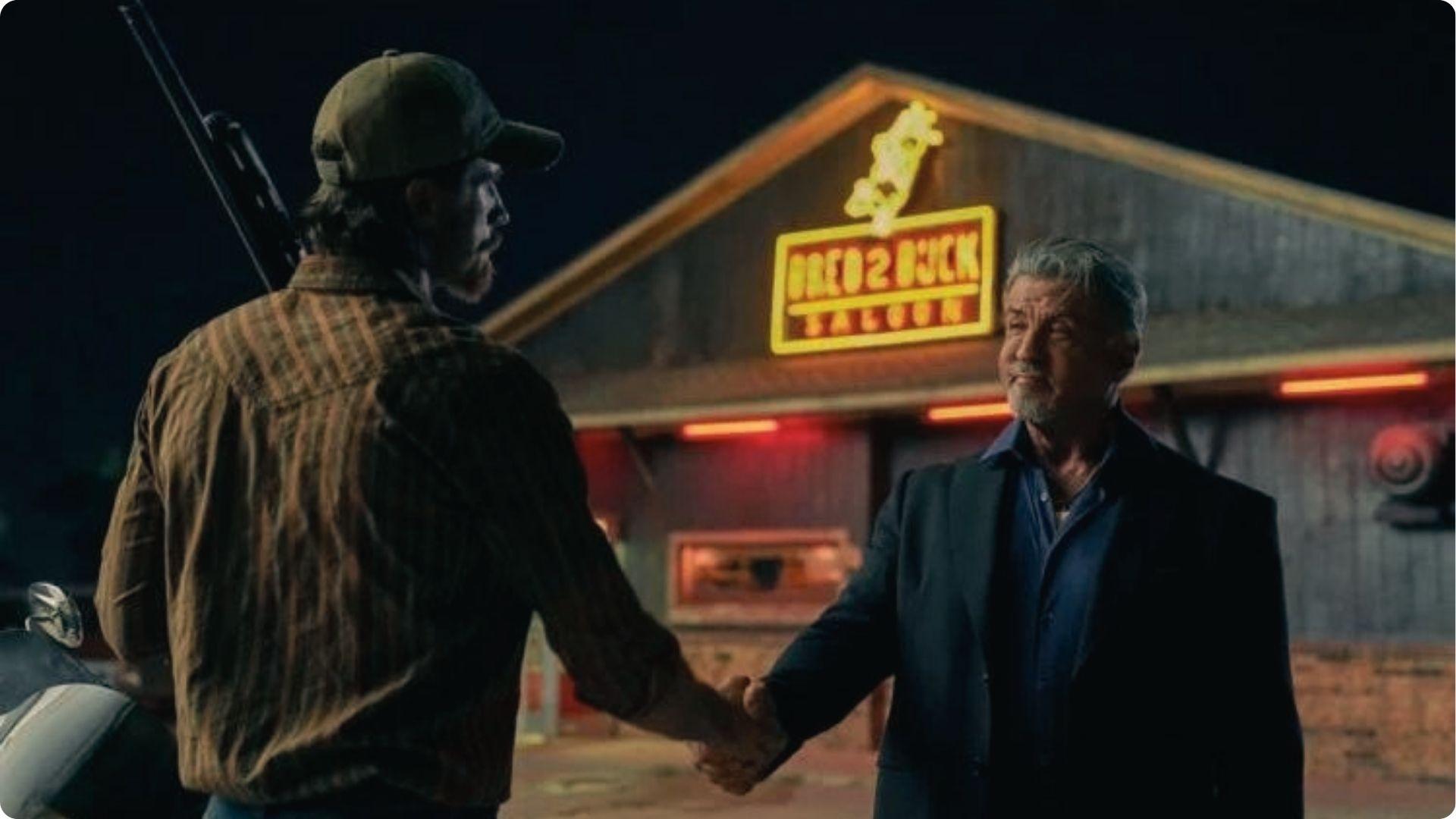 Sylvester Stallone shakes hands with a man holding a rifle outside BRED2BUCK Saloon at night, a Tulsa King filming location.