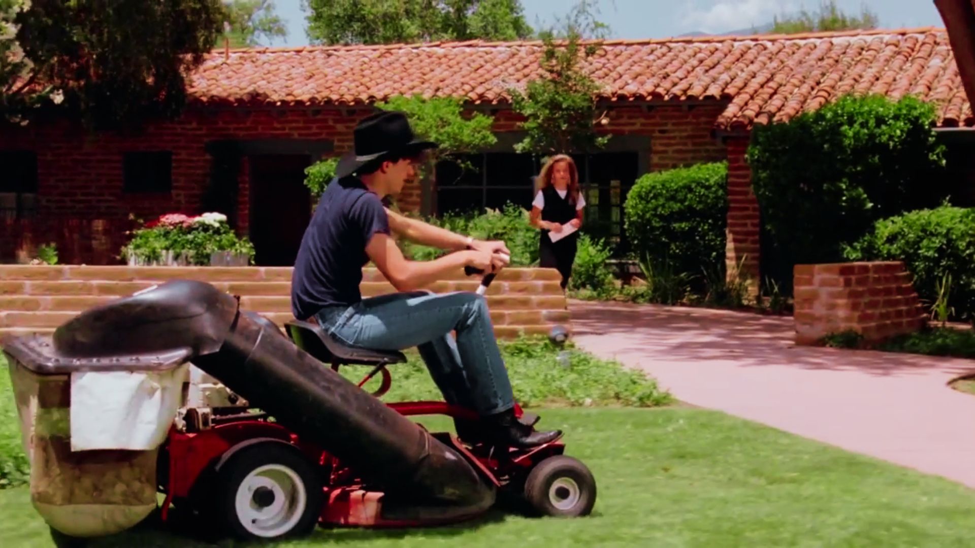 Teen in cowboy hat rides a lawn mower in front of a brick house while a girl watches.