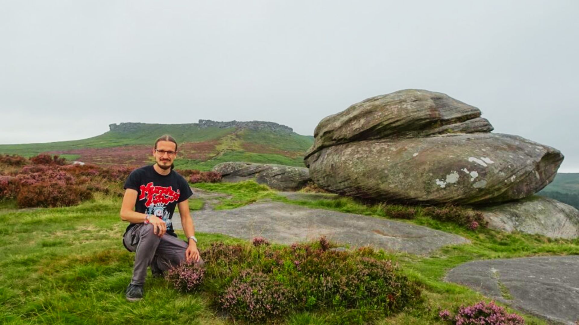 A person kneels among heather and gritstone boulders with Carl Wark hillfort visible in the distance.