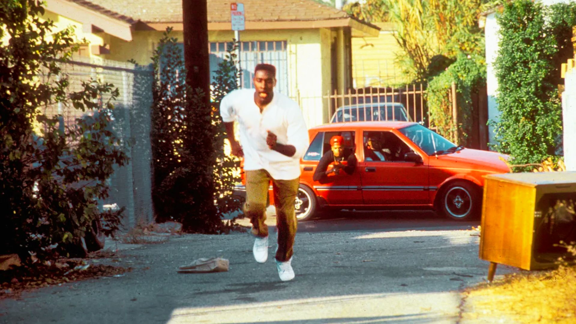 A young man in a white shirt and khaki pants sprints down a South Central Los Angeles alley in daylight, looking over his shoulder.