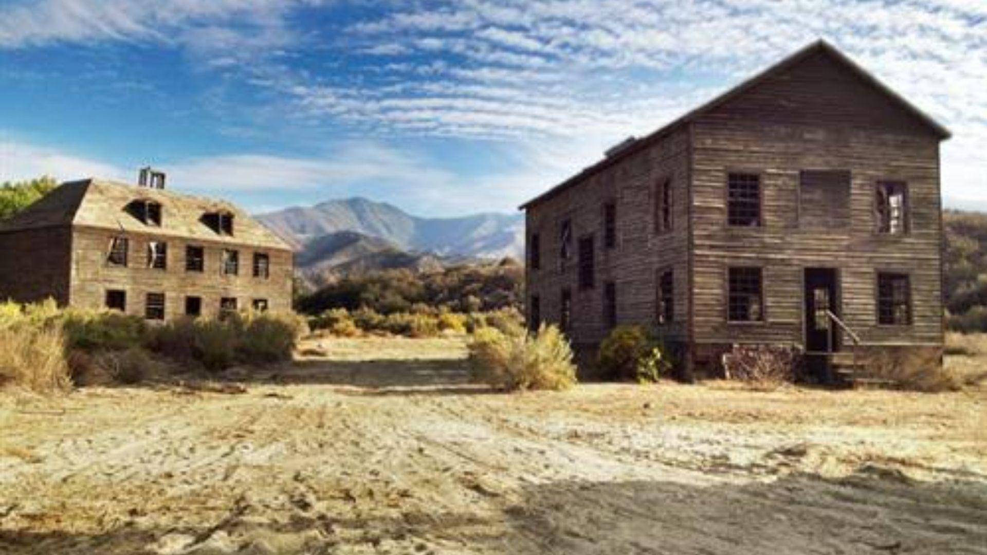 Still from NOPE. Two abandoned two-story wooden buildings on dusty lot beneath cloud-streaked mountains.