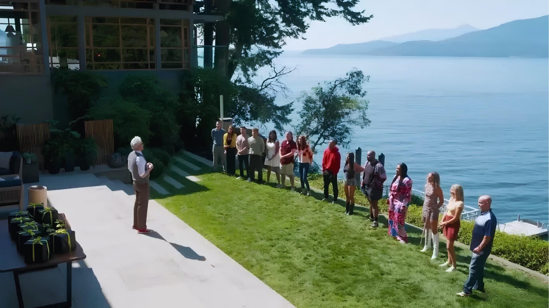 Host in argyle sweater vest, yellow tie, and red sneakers addresses line of 14 contestants on grassy lawn overlooking ocean, with black gift boxes on table.