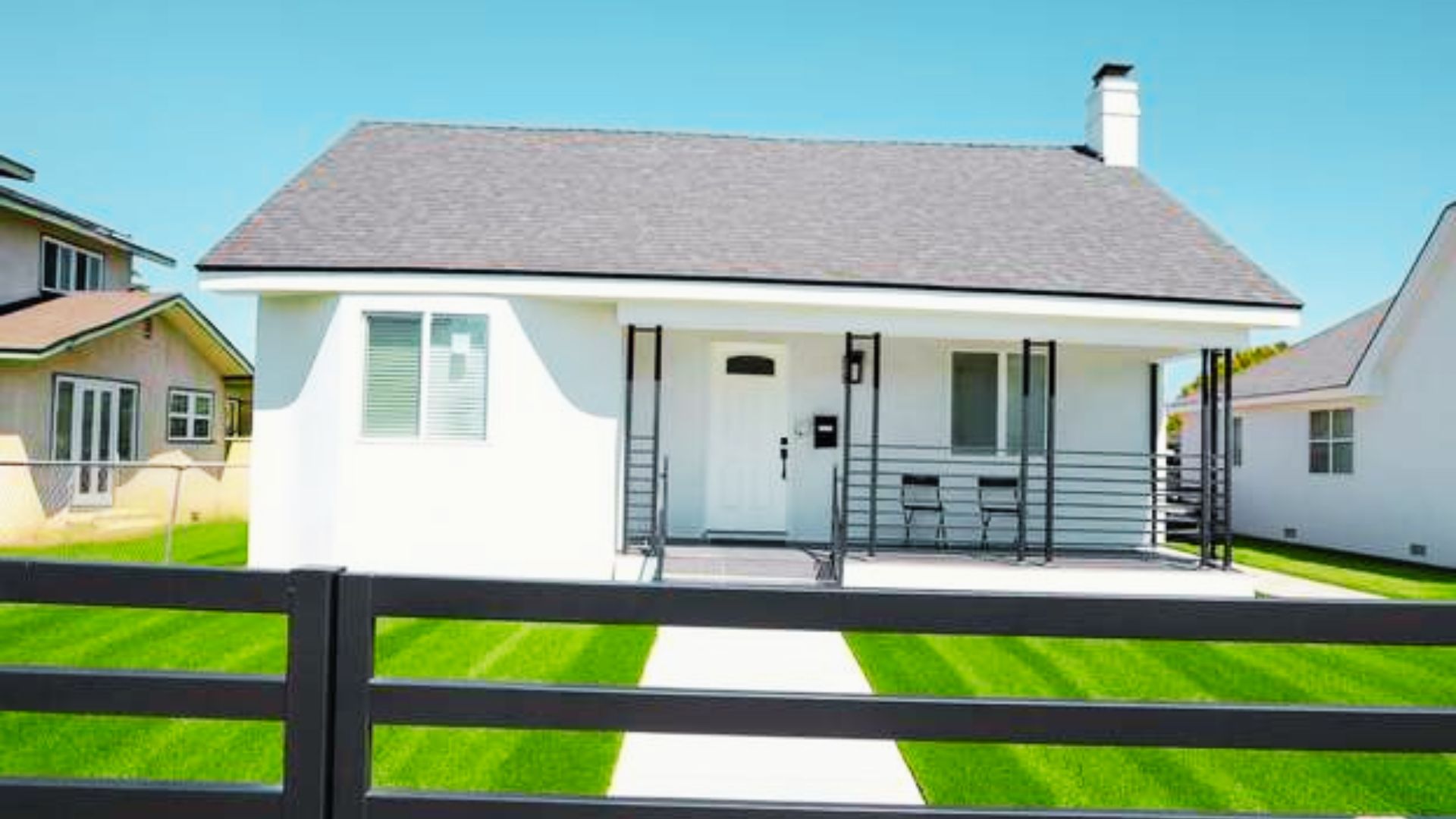 White single-story house with black railings, bright lawn, behind a black fence.