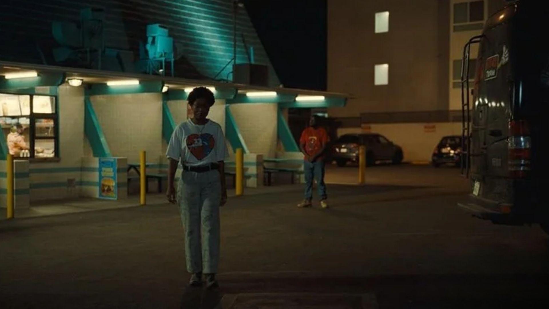 Still from NOPE. Young Jupe in white graphic tee crosses teal-lit fast-food parking lot at night.