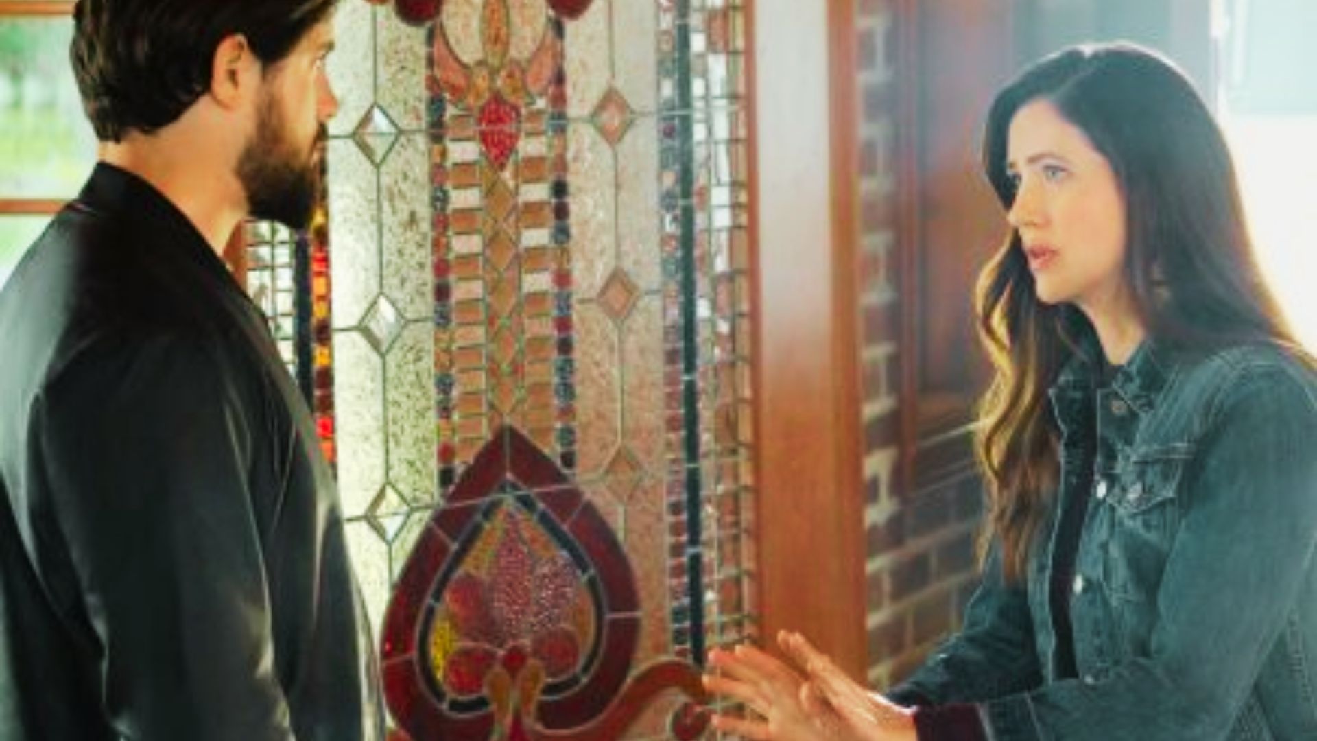 A bearded man and woman face each other by ornate stained glass inside the historic house.
