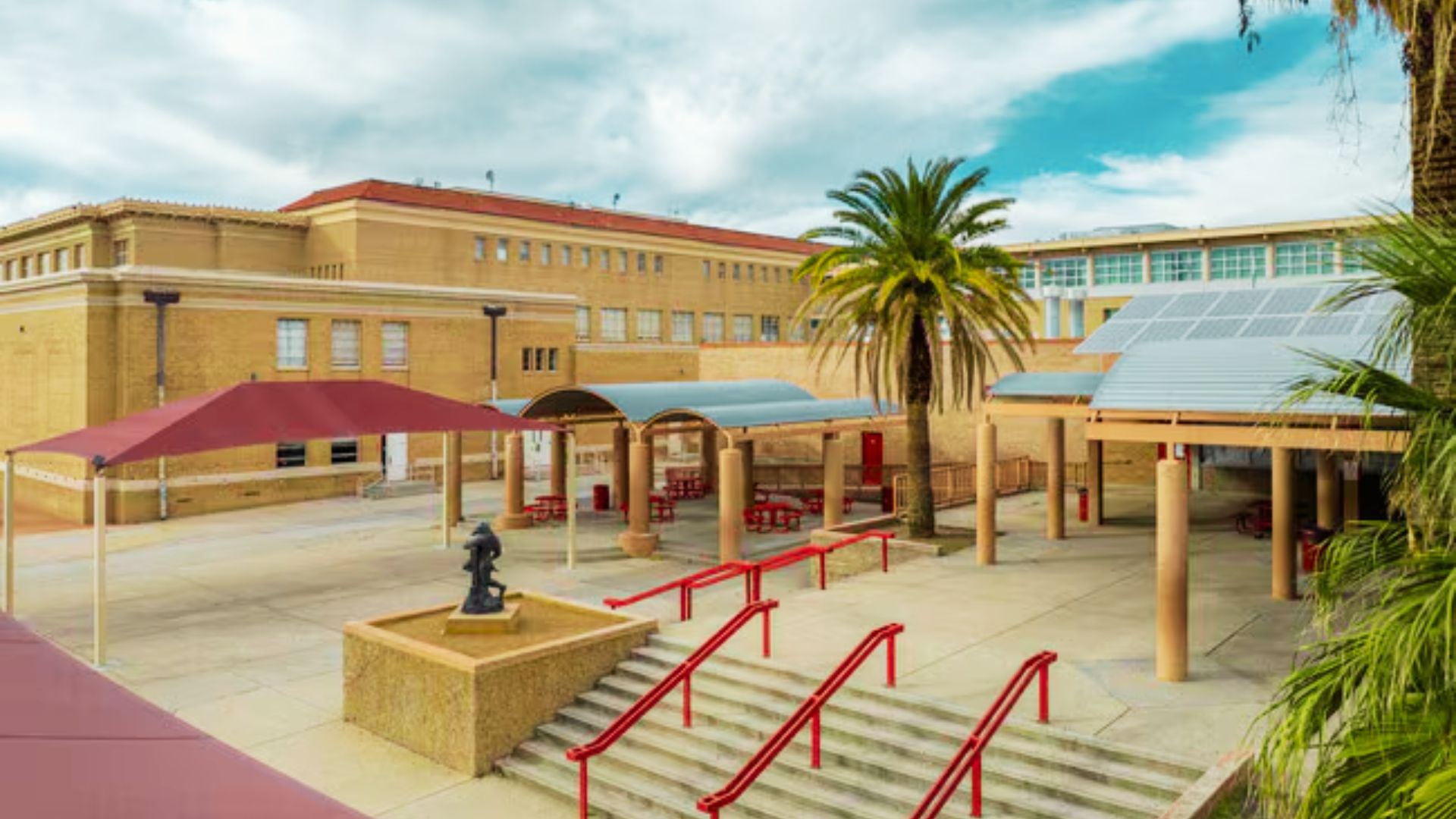 Tucson High School courtyard with red shade structures, palm trees, and statues by steps.