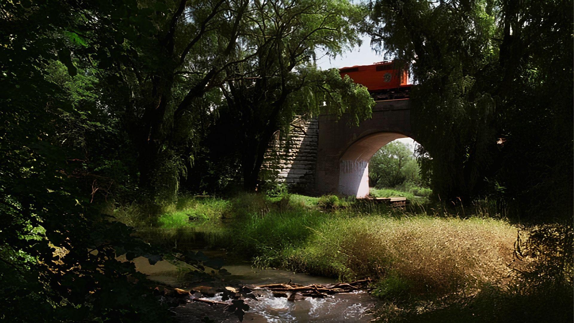 A train crosses a bridge over a flowing stream, surrounded by lush greenery and a clear blue sky.