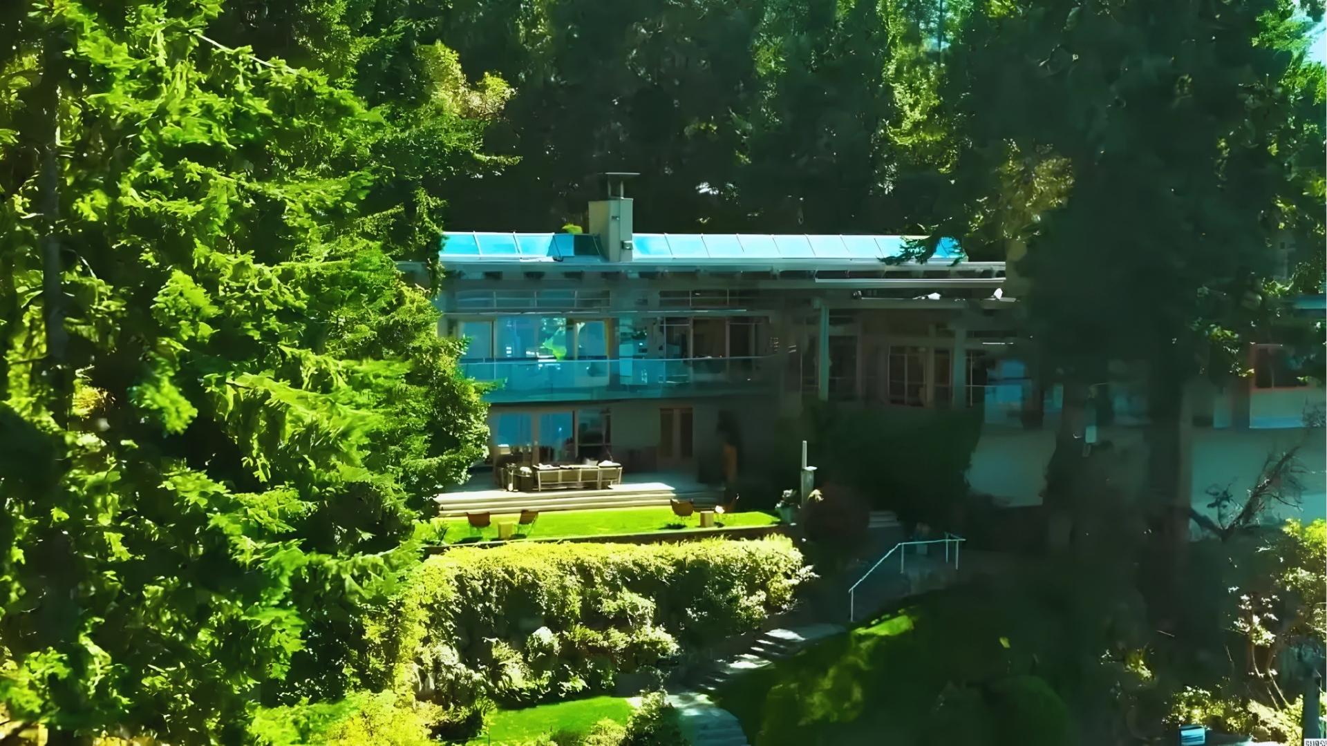 Daytime view of modern glass house with tiered lawns, patio furniture, and blue roof, surrounded by lush green trees and landscaped gardens.