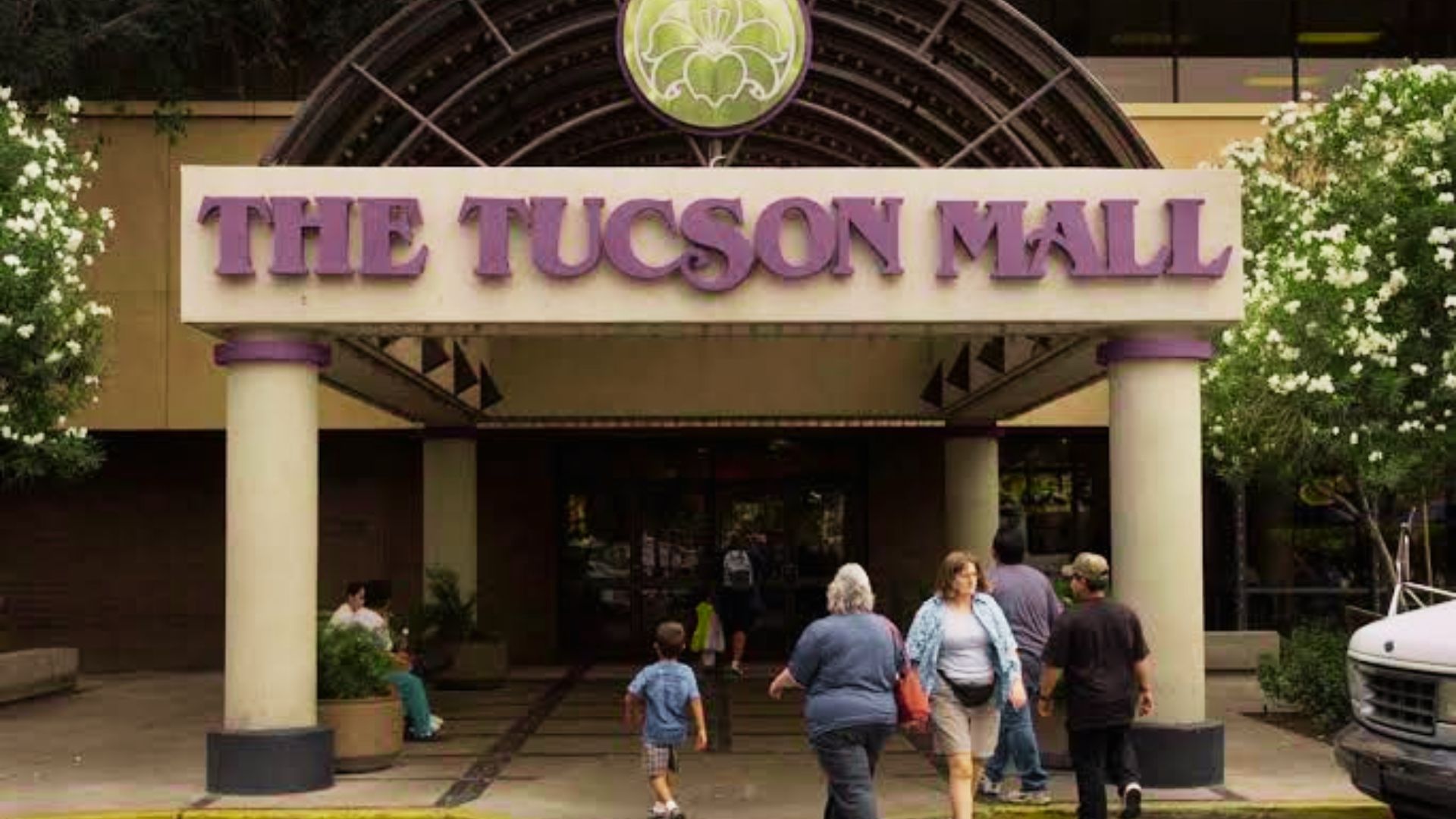 Shoppers walk into The Tucson Mall entrance with purple lettering and arched facade.