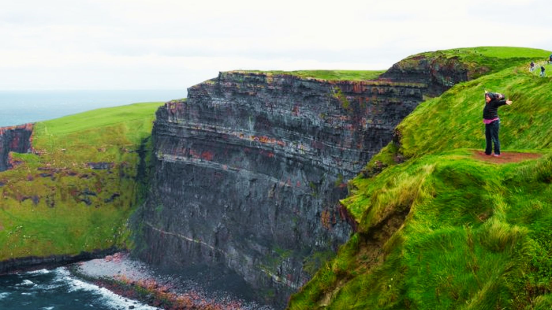 A person stands with arms outstretched on the grassy edge of the Cliffs of Moher overlooking the Atlantic.