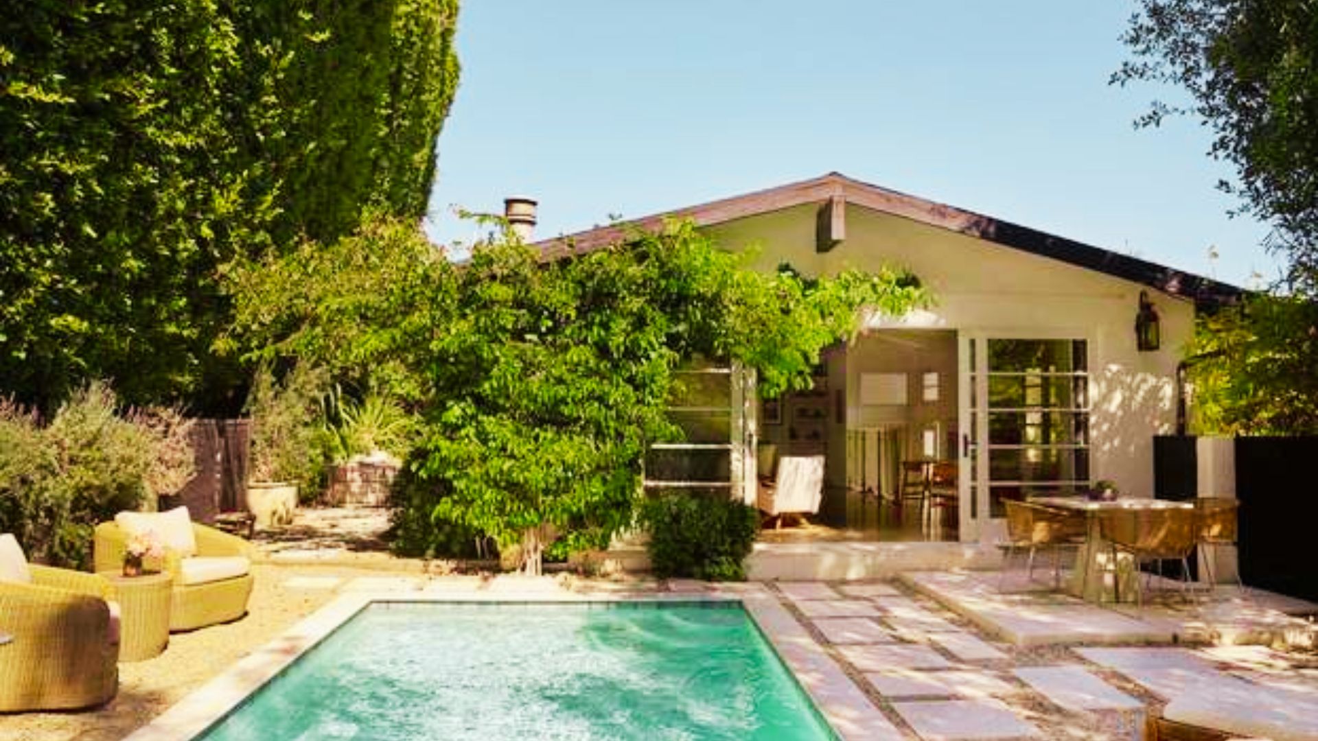 Sunny backyard with swimming pool, patio, wicker chairs, and glass doors leading into a white house.