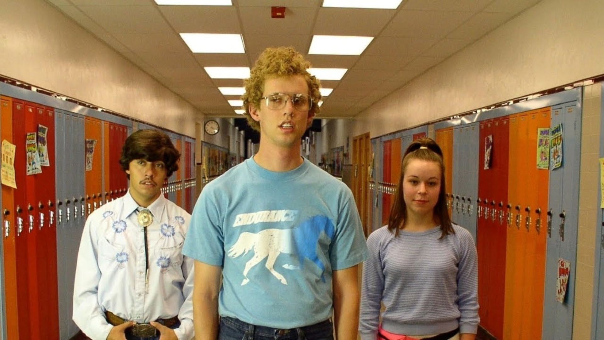 Three individuals stand in a school hallway lined with lockers, discussing the film's cult classic status.