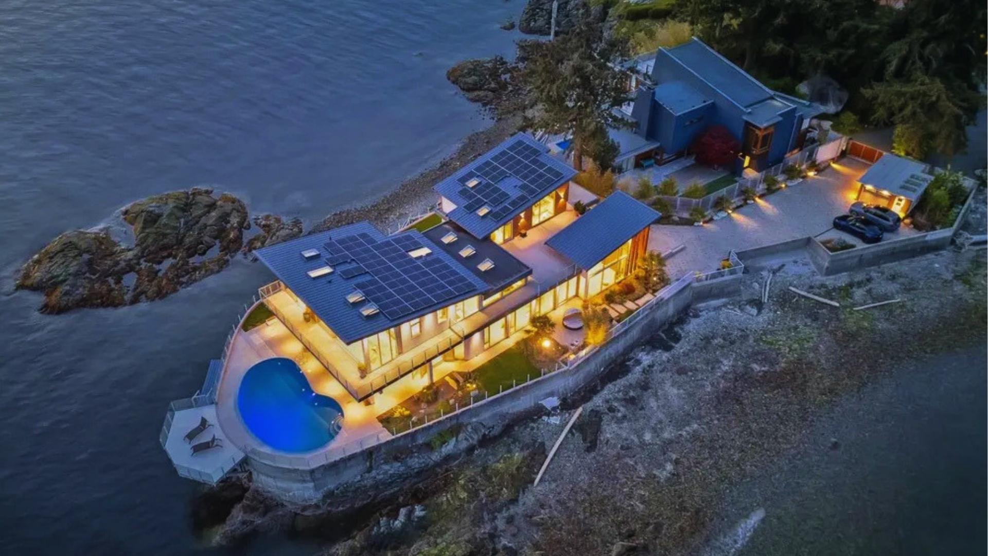 Aerial dusk view of illuminated modern waterfront mansion with solar panels and kidney-shaped pool, perched on rocky ocean shoreline with cars parked.