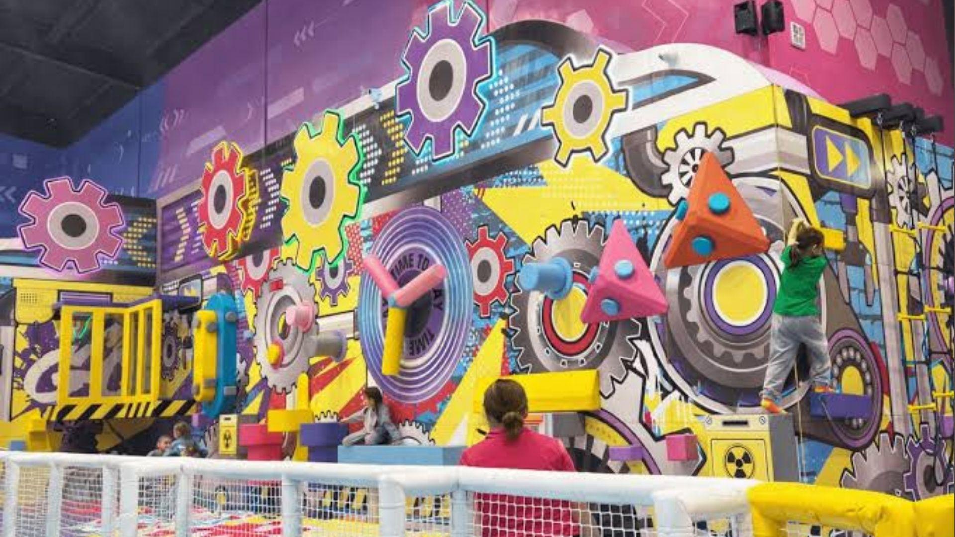 Kids climbing colorful gear-themed wall with giant clock and 3D shapes at indoor play center