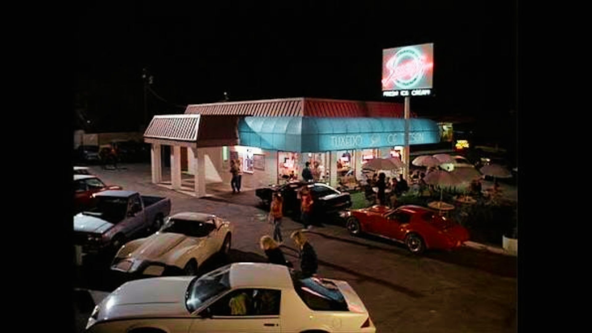 Night scene of Tuxedo Tucson diner with neon sign, vintage cars, and teens gathering outside. 