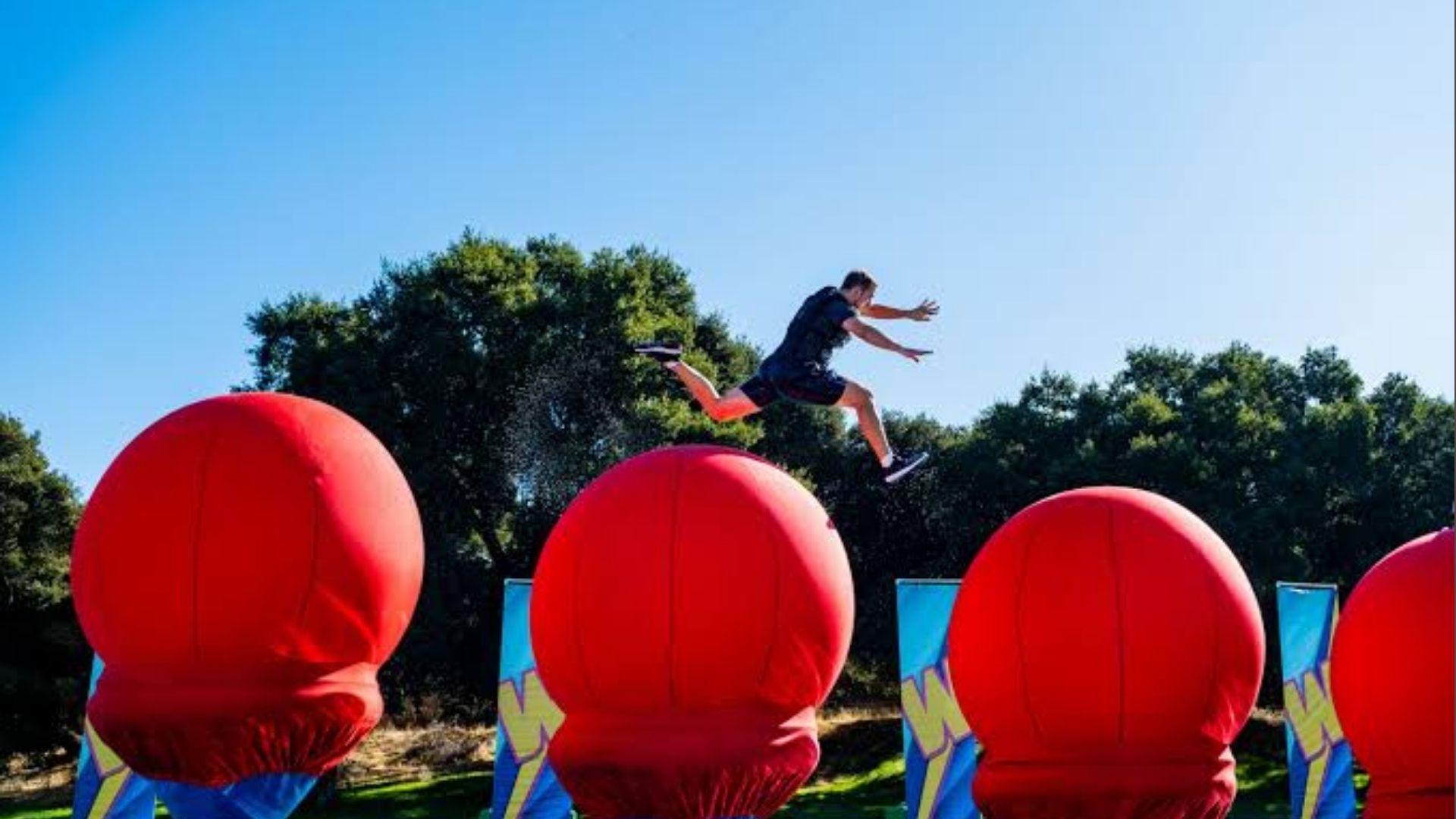 Contestant leaping between giant red inflatable balls on Wipeout obstacle course