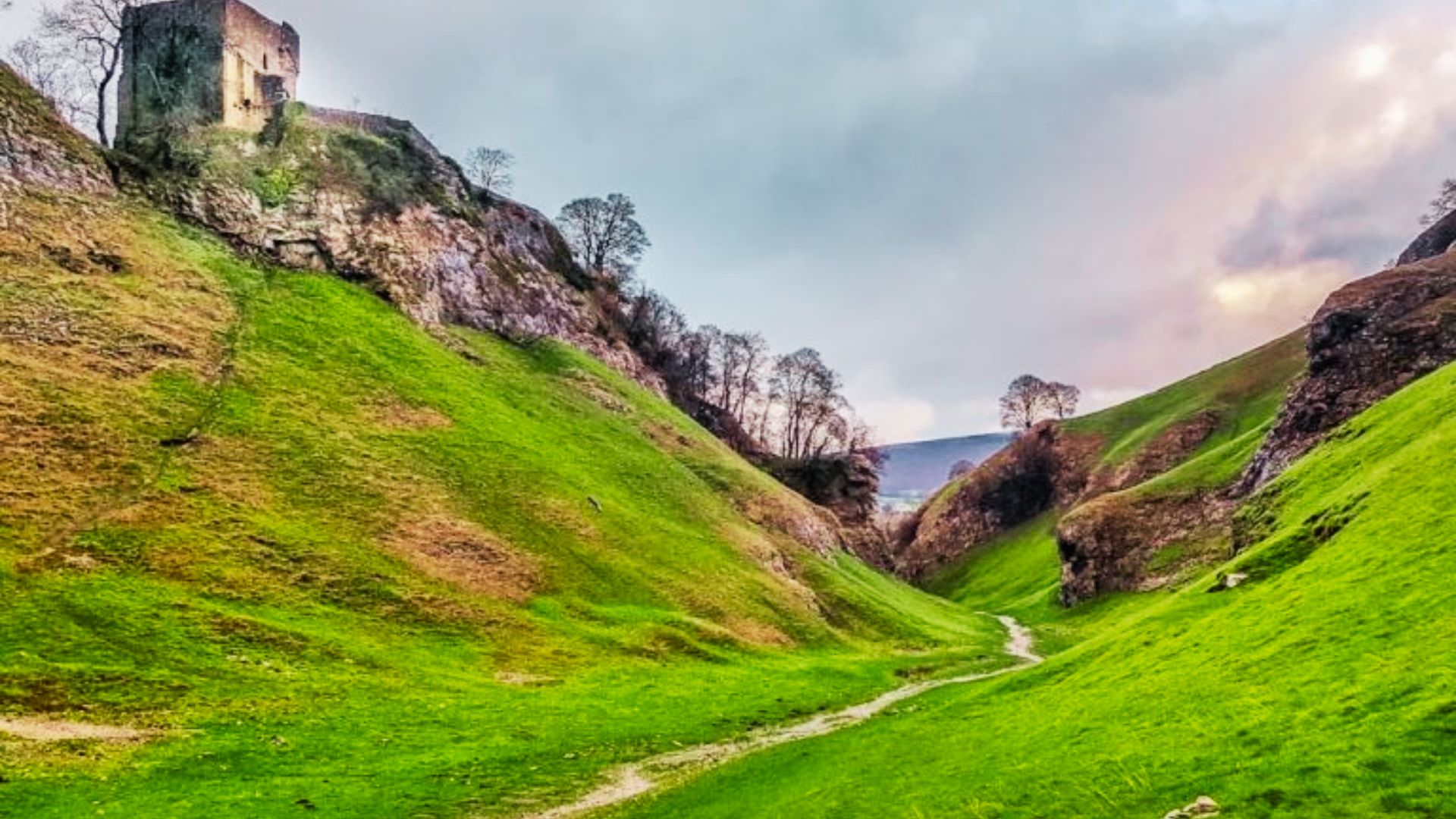 A narrow limestone valley path winds through green slopes below the ruins of Peveril Castle.
