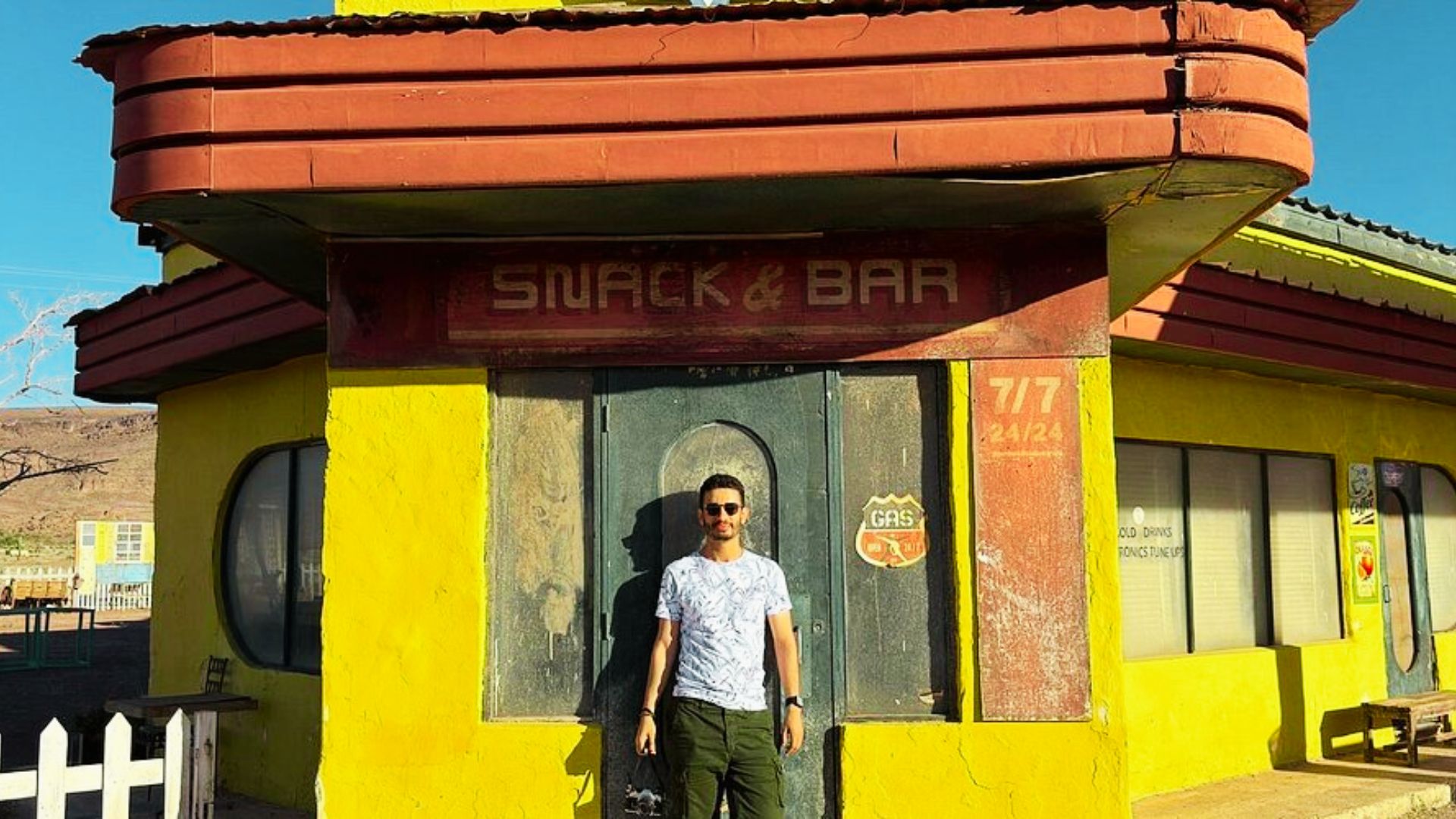 Man stands in front of a bright yellow Snack & Bar building with Gas signs in the desert.