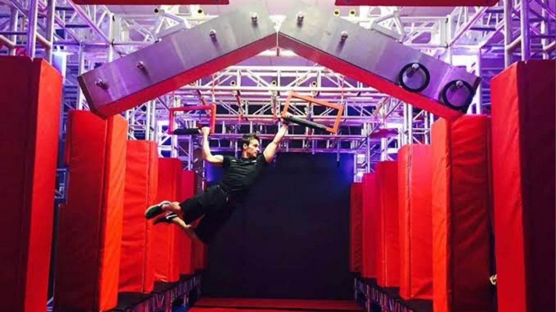 Athlete swinging on hanging grips in indoor ninja obstacle course with red pads and metal trusses