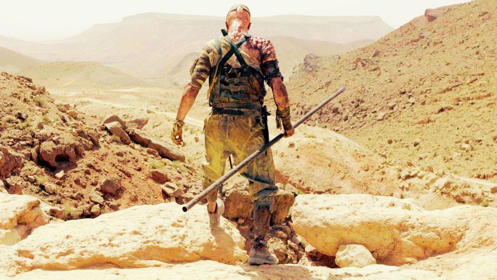 A disfigured mutant stands with a metal pipe overlooking a vast desert canyon.