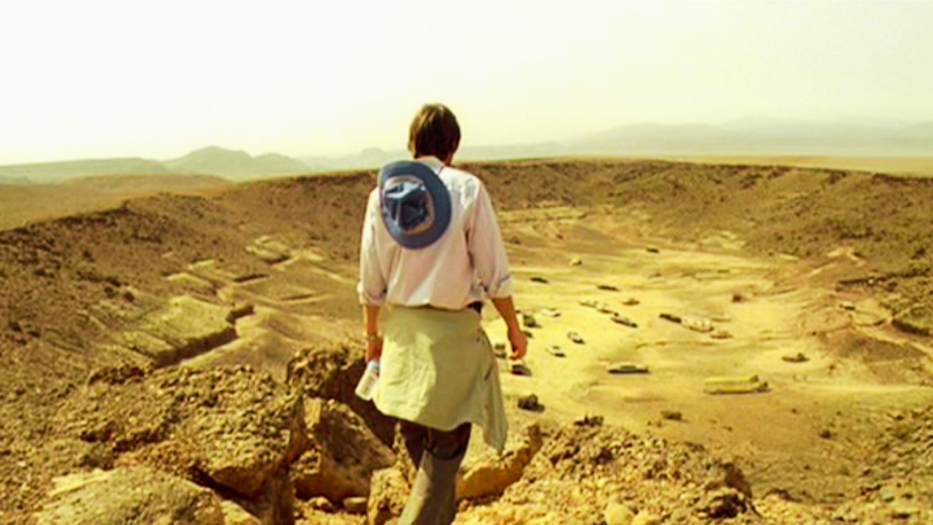 Person overlooking a sunbaked canyon filled with abandoned vehicles from a rocky cliff.