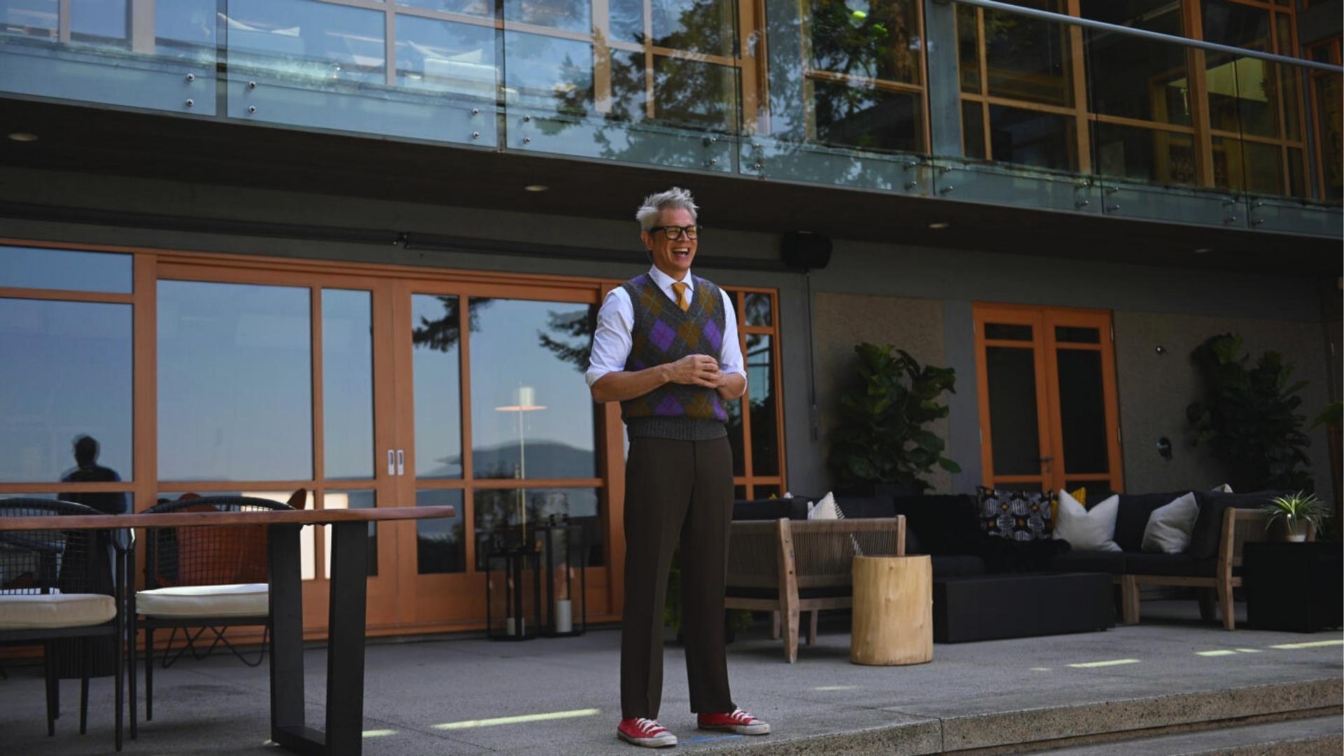 Host with gray hair, black glasses, argyle sweater vest, yellow tie, and red sneakers laughs while standing on modern patio outside glass house.
