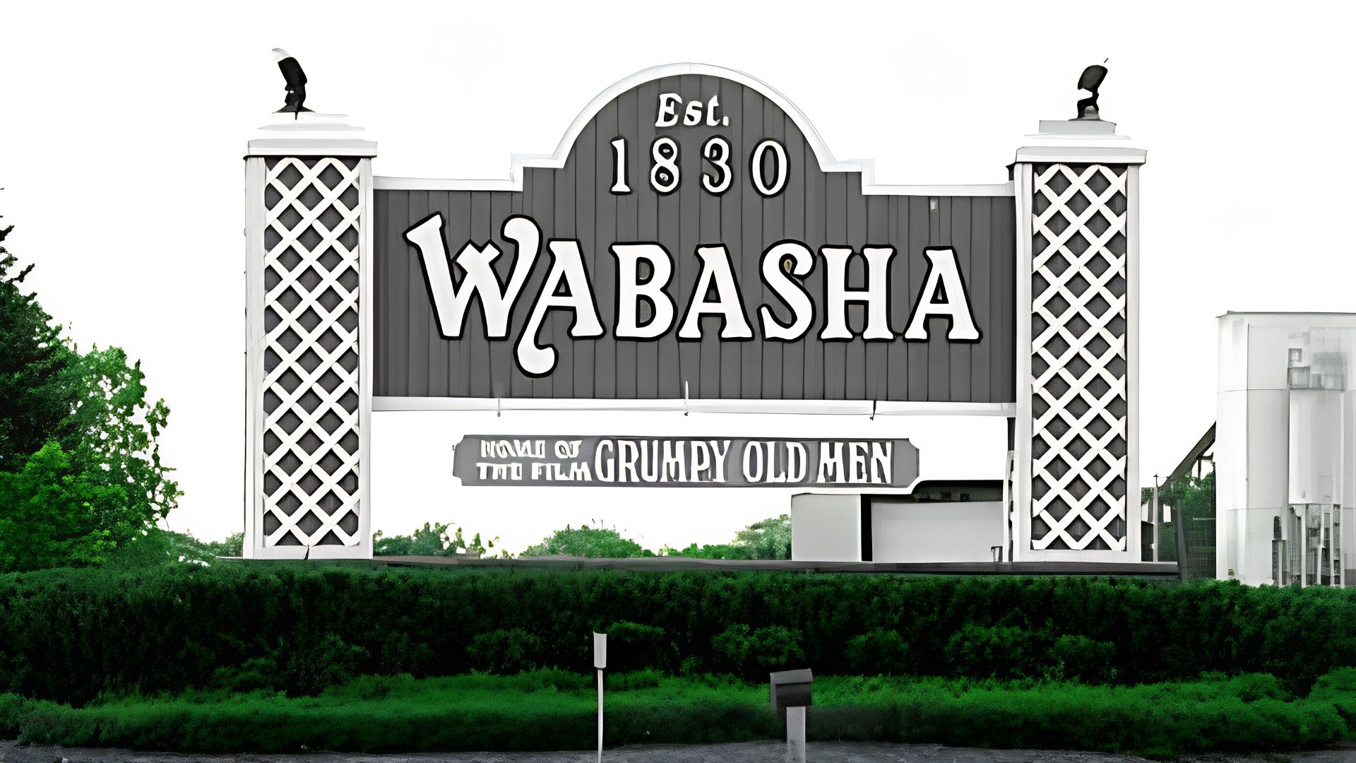 Welcome sign for Wabasha, Minnesota, featuring the town name, founding year 1830, and a reference to Grumpy Old Men, surrounded by greenery.