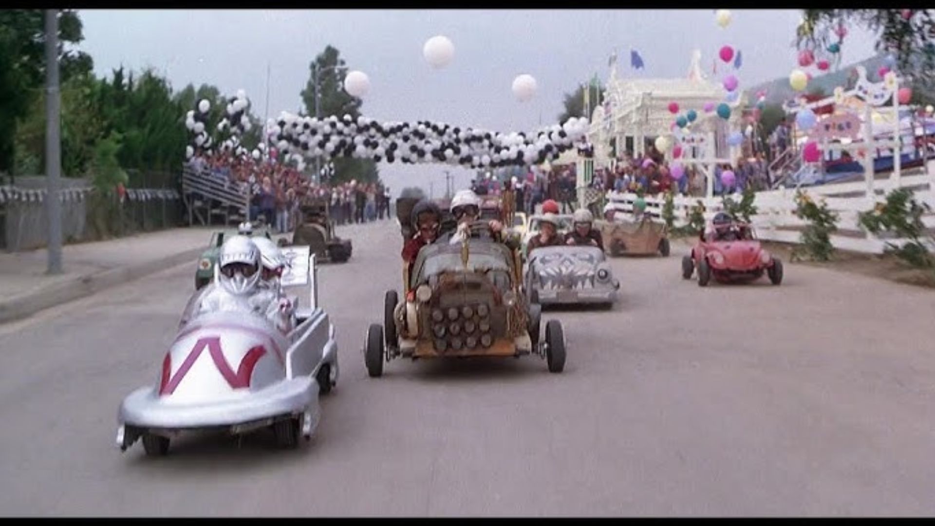 Little Rascals 1994 soapbox derby race scene with kids driving homemade cars down a street decorated with balloons and crowd