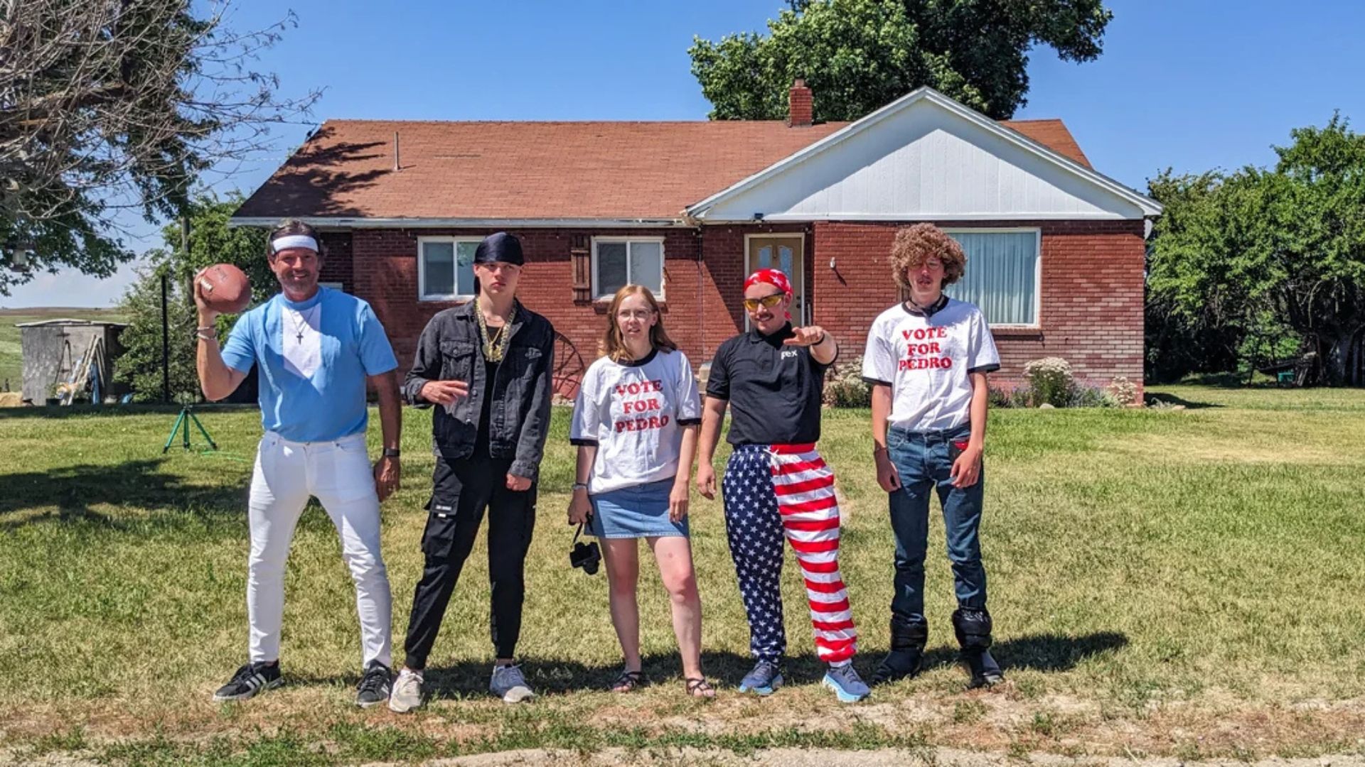A group of four people poses in front of a house featuring American flags, highlighting a scene from the filming of Napoleon Dynamite.