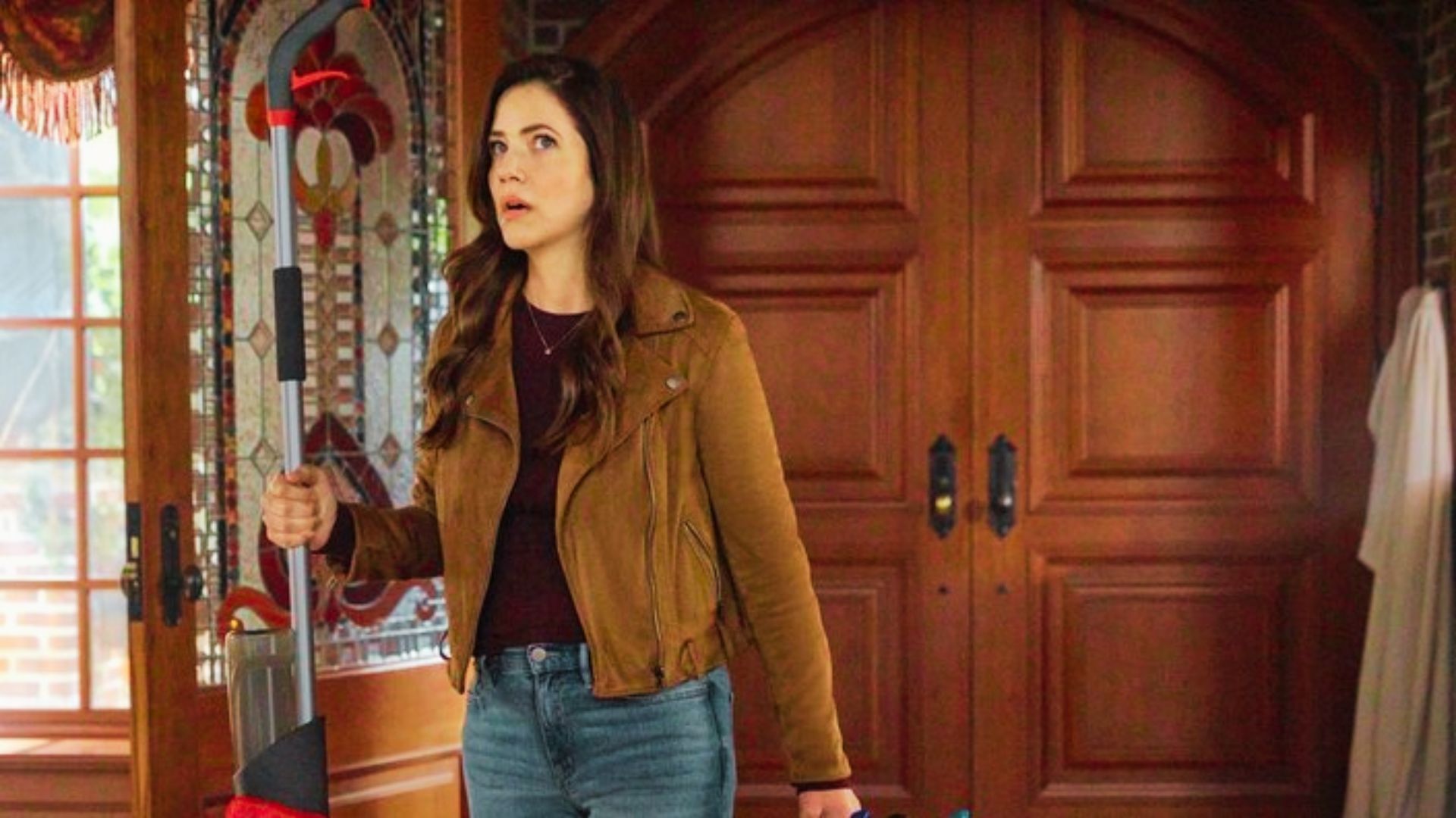 A startled woman grips a mop inside a grand entryway with stained glass and wood doors.