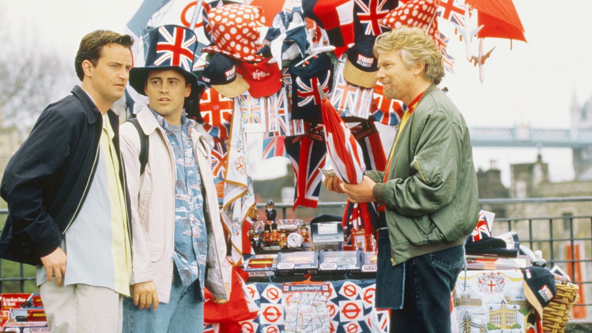 Three men stand in front of a display of British flags during the filming of a Friends TV series episode.