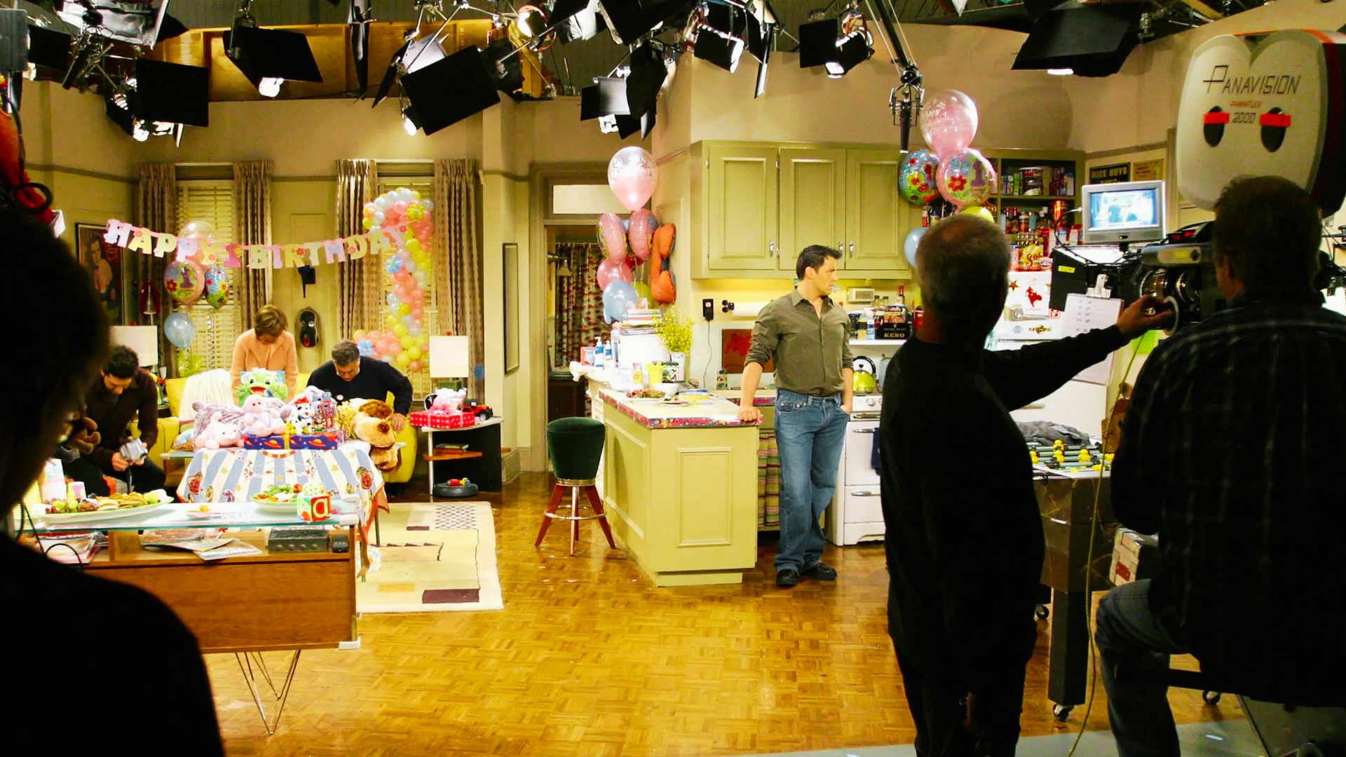 A kitchen scene featuring friends filming, with a television present, highlighting a moment from a popular TV series.