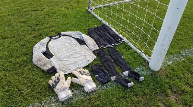 Goalkeeper gloves and clothing laid out on turf near a football goal